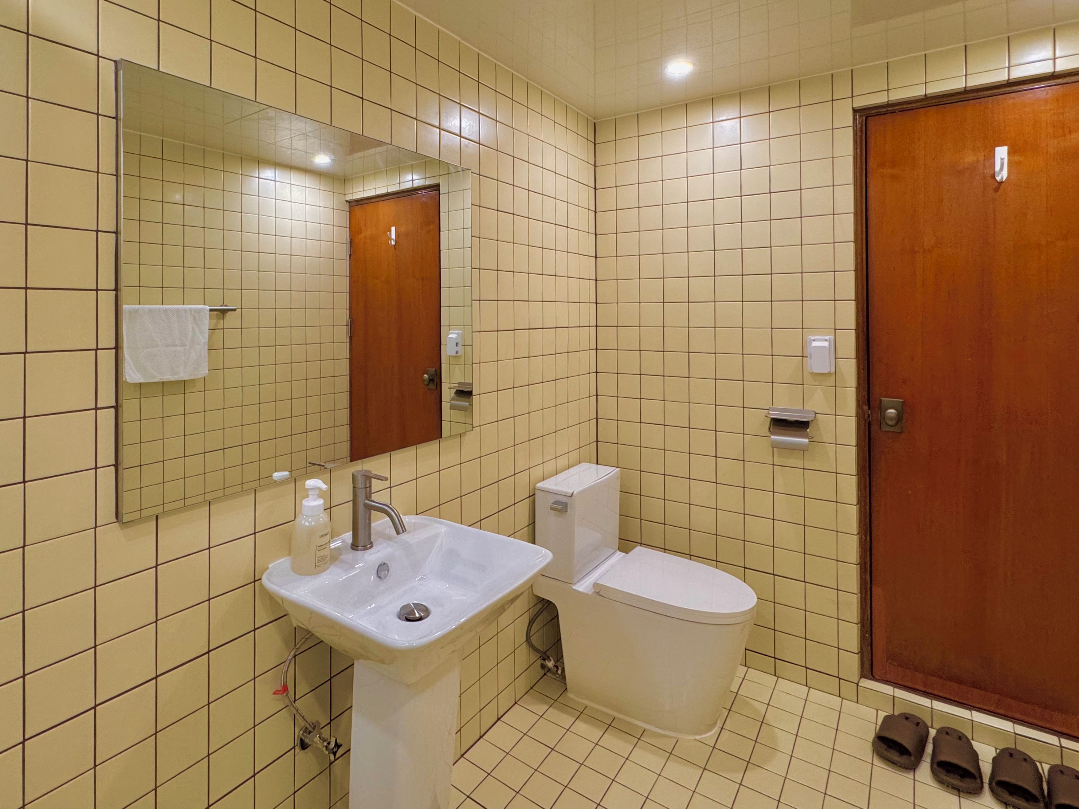 A bathroom with a sink, toilet, and mirror. There are slippers on the floor.