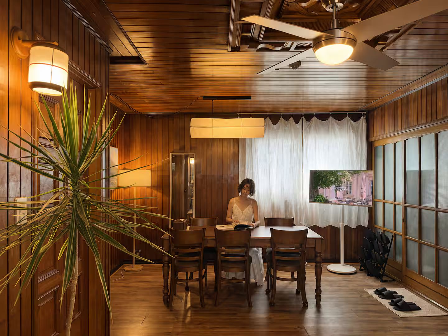 A room with wooden paneling, a dining table with chairs, a large plant, and a ceiling fan.