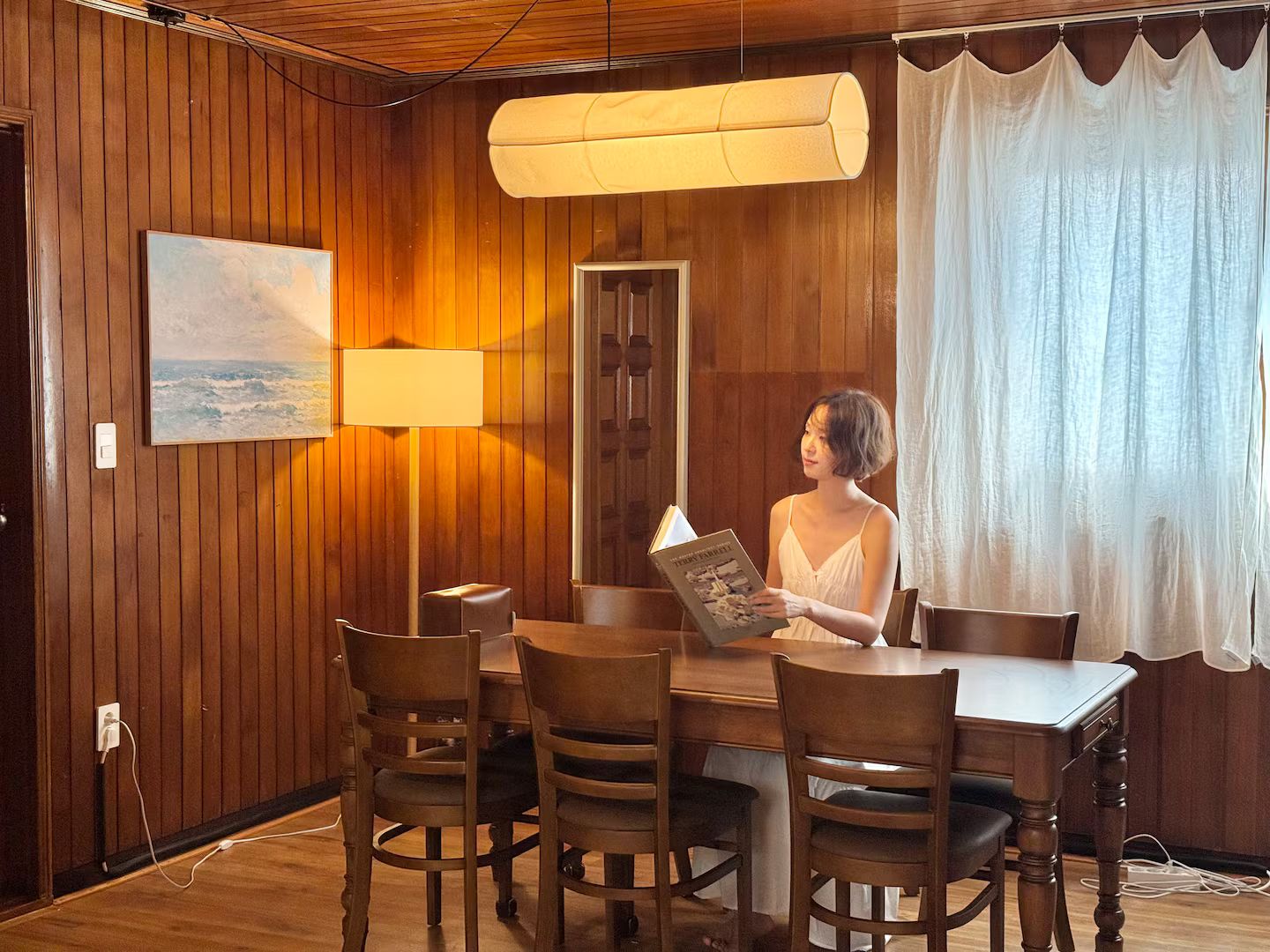 A wooden-paneled room with a dining table, chairs, a lamp, a painting, and a person reading a book.