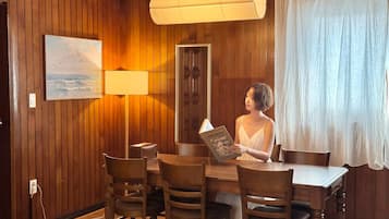 A wooden-paneled room with a dining table, chairs, a lamp, a painting, and a person reading a book.