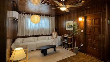 A room with a wooden ceiling, a ceiling fan, a white sofa, a black bench, a lamp, a potted plant, and a door with a framed picture.