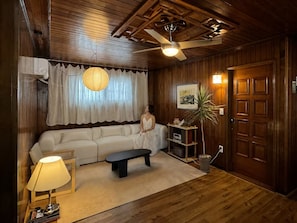 Ett rum med ett träpanelat tak, en takfläkt, en vit soffa, en svart bänk, en lampa, en krukväxt och en dörr med en inramad tavla.