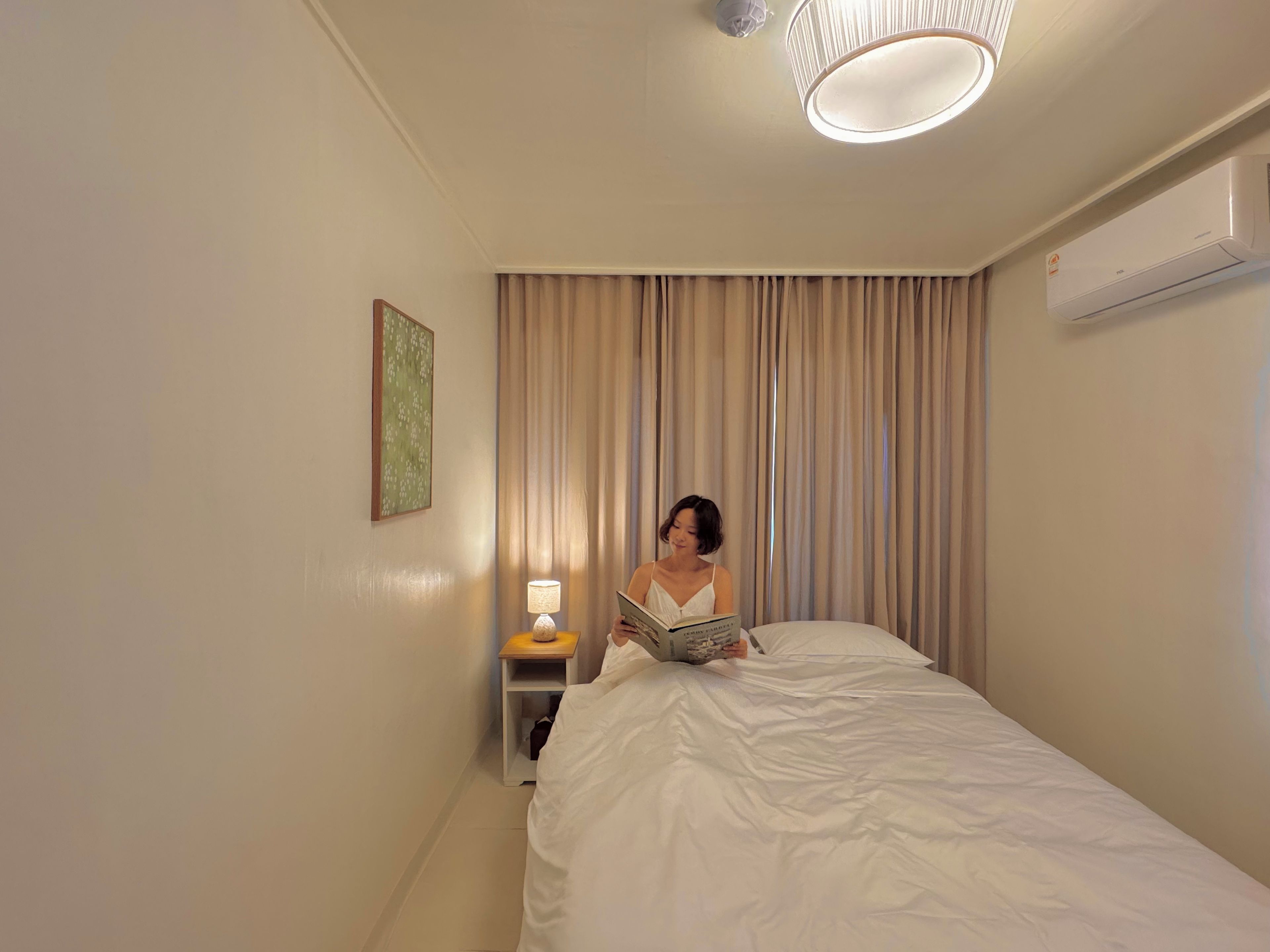 A hotel room with a bed, a bedside lamp, a wall-mounted air conditioner, and a window with curtains.