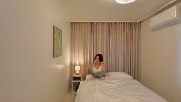 A hotel room with a bed, a bedside lamp, a wall-mounted air conditioner, and a window with curtains.