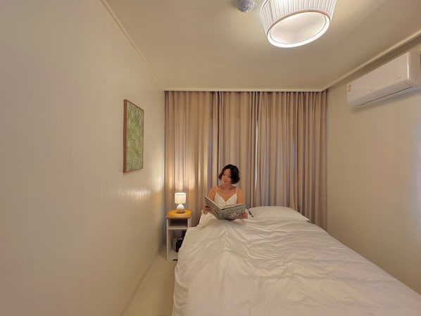 A hotel room with a bed, a bedside lamp, a wall-mounted air conditioner, and a window with curtains.