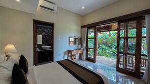 A hotel room with a bed, a desk, a mirror, and a view of a garden through large windows.