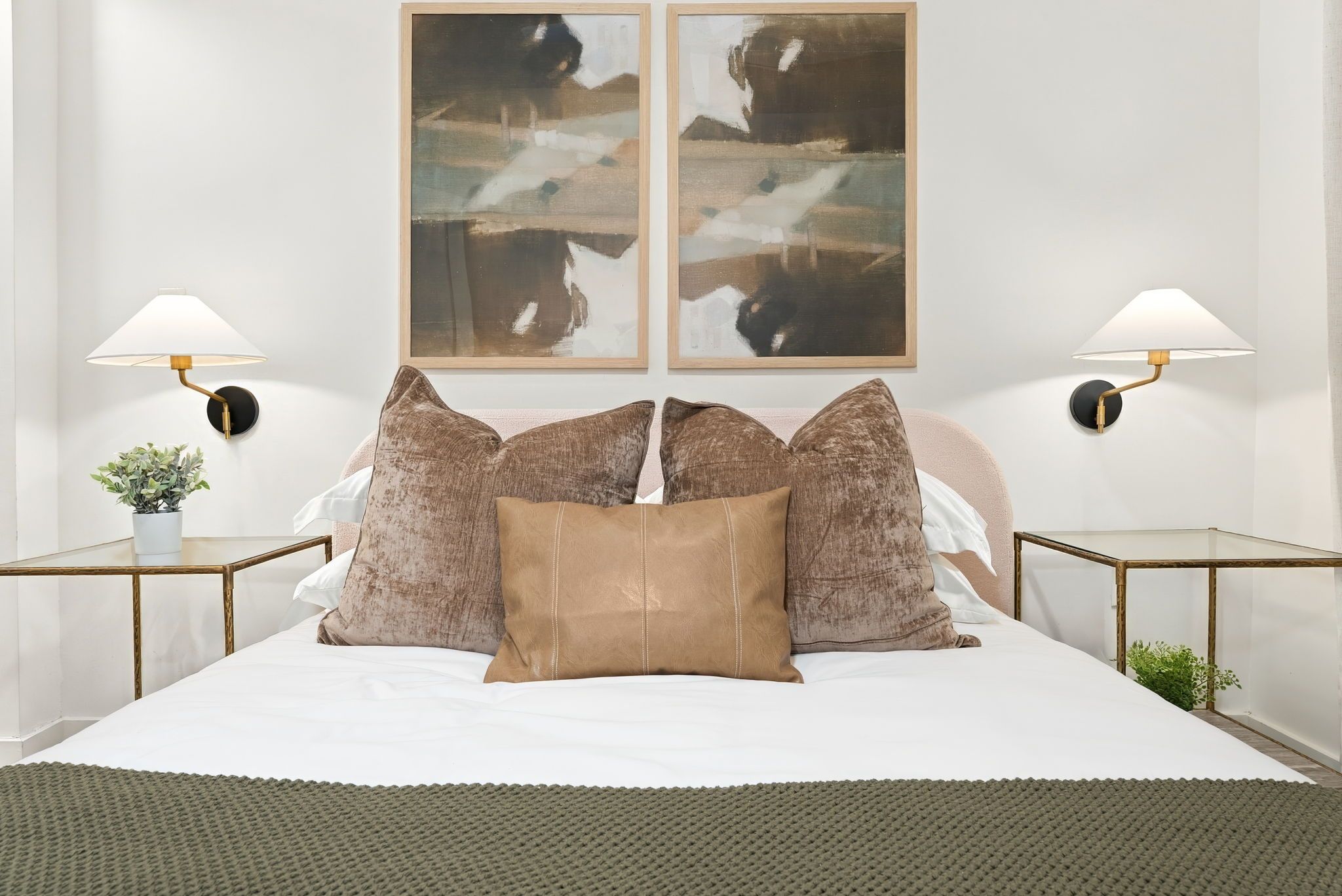 A bed with a white sheet, two brown pillows, and a green knitted throw. There are two wall-mounted lamps with round bases and a plant on a side table.