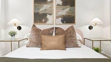 A bed with a white sheet, two brown pillows, and a green knitted throw. There are two wall-mounted lamps with round bases and a plant on a side table.