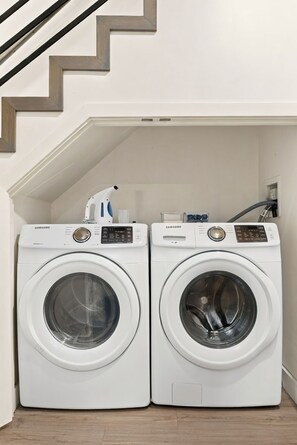Deux lave-linge Samsung à chargement frontal, de couleur blanche, se trouvent dans une buanderie. On aperçoit, à l’arrière-plan, un escalier en bois.