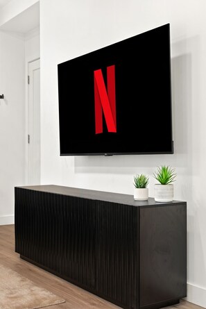 A modern living room with a large flat-screen TV mounted on the wall, a dark wooden console table, and two potted plants. - New Lux Boston Apt - Free Parking - Near Fenway (Boston)