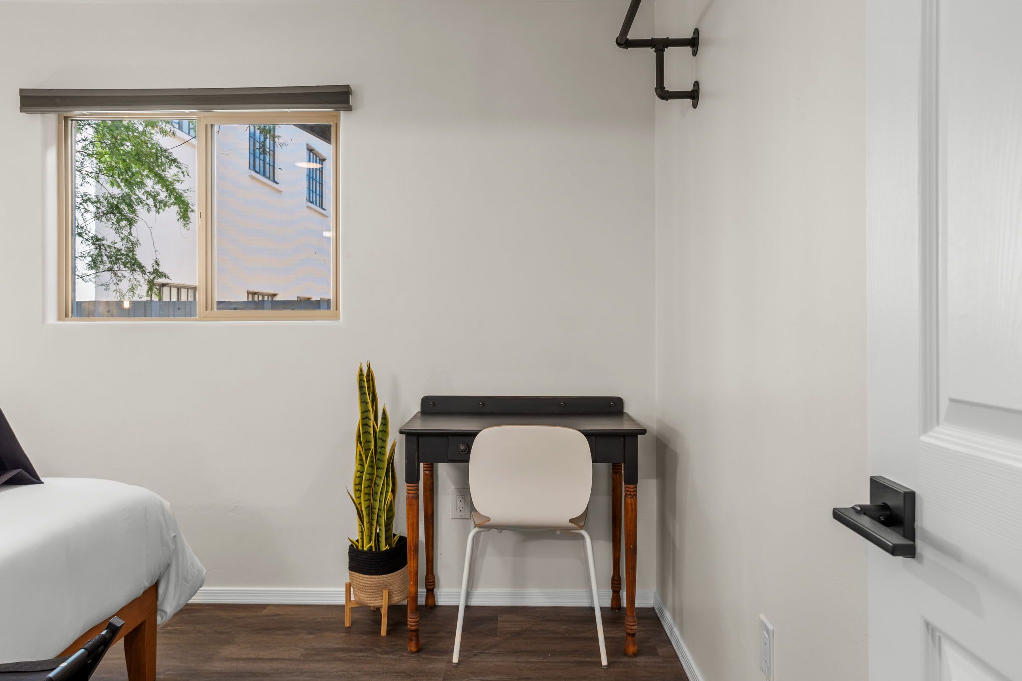 A bedroom with a bed, a desk, a chair, a potted plant, and a window with a view of buildings.