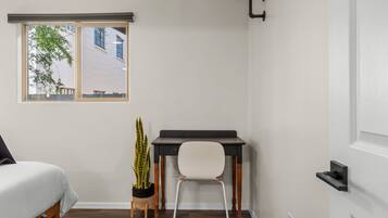 A bedroom with a bed, a desk, a chair, a potted plant, and a window with a view of buildings.