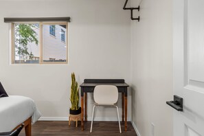 Une chambre à coucher comprenant un lit, un bureau, une chaise, une plante en pot et une fenêtre donnant sur des immeubles.