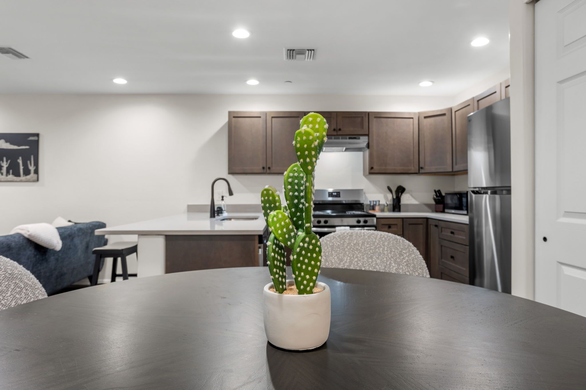 A modern kitchen with wooden cabinets, a stainless steel refrigerator, and a dining table with a cactus centerpiece.