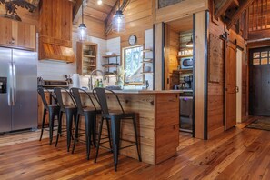 A modern kitchen with a central island, bar stools, wooden cabinetry, and a stainless steel refrigerator. - Castle Rock Luxury Cabin with Smoky Mt Views (Bryson City)