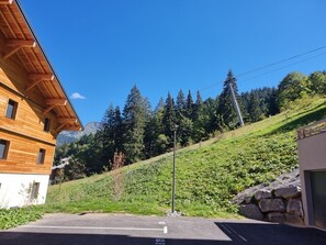 Ein modernes Gebäude mit Holzfassade, einem gepflasterten Zugang und einer hügeligen Landschaft mit Bäumen und blauem Himmel.