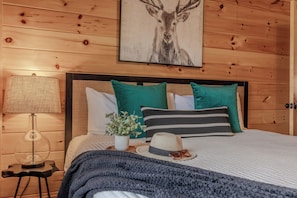 A wooden-paneled bedroom with a bed, pillows, a lamp, a hat, sunglasses, and a framed picture of a deer. - 84° Indoor Pool, 75” TV, Hot Tub, Air Hockey, WIFI, Arcade, A+ Location (Pigeon Forge)