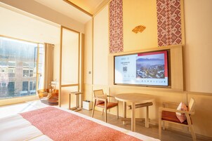Habitación de hotel con una ventana grande, una cama, un escritorio, una silla y un televisor mostrando un paisaje urbano.
