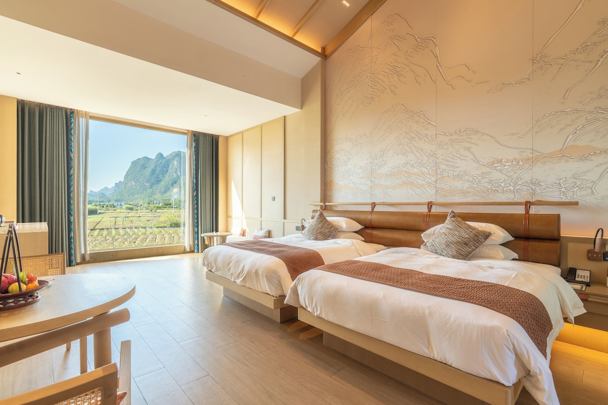 A hotel room with two beds, a dining table, and a large window with a view of mountains.