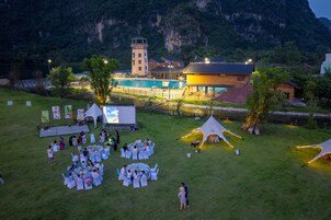 An outdoor event with round tables and chairs, a stage, and tents set up in a grassy area with a mountain backdrop.