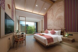 A modern hotel room with a large bed, a dining table, and a view of greenery.