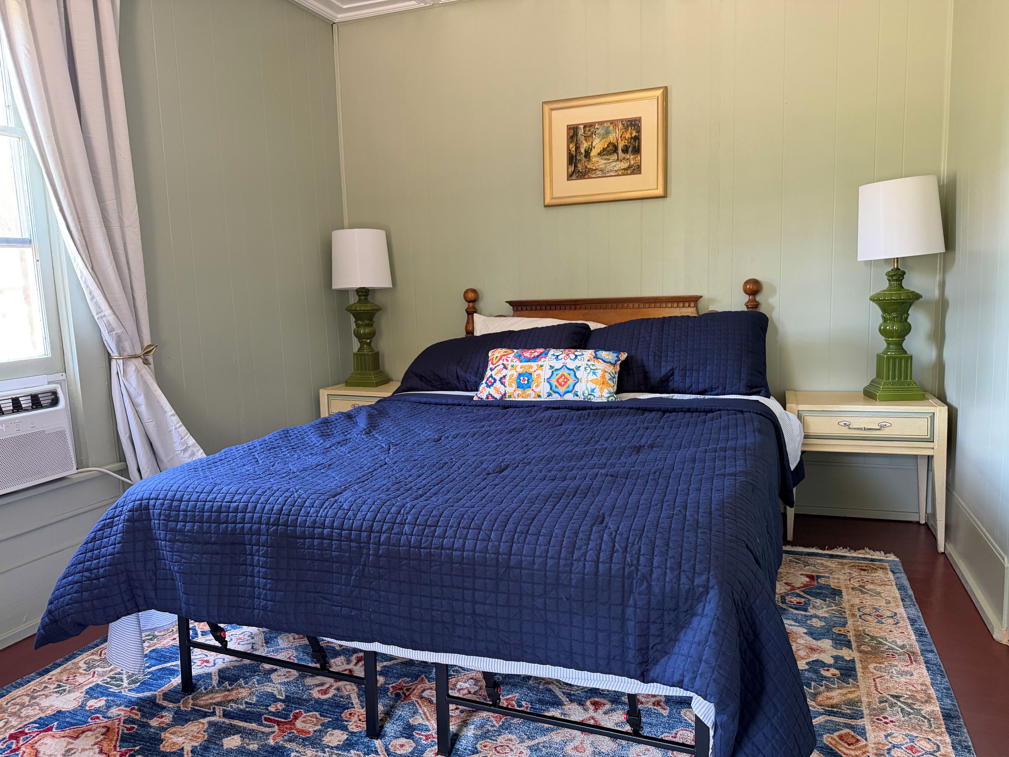 A neatly made bed with a blue quilt, two bedside lamps, a patterned pillow, a framed picture on the wall, and a window with curtains.