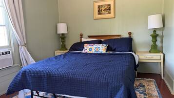 A neatly made bed with a blue quilt, two bedside lamps, a patterned pillow, a framed picture on the wall, and a window with curtains.