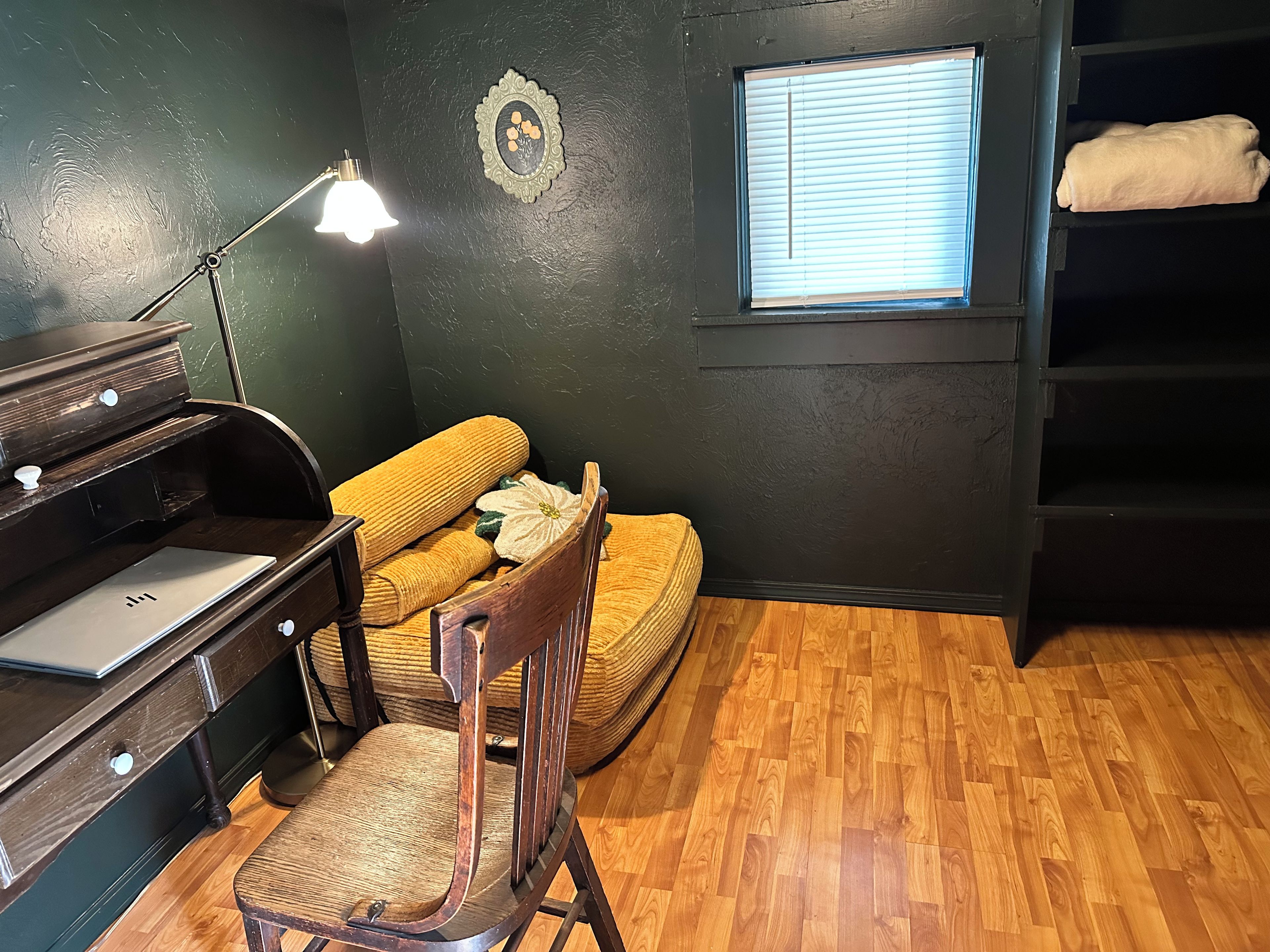 A room with a wooden floor, a desk with a lamp, a chair, a window with blinds, and a shelf with a blanket.