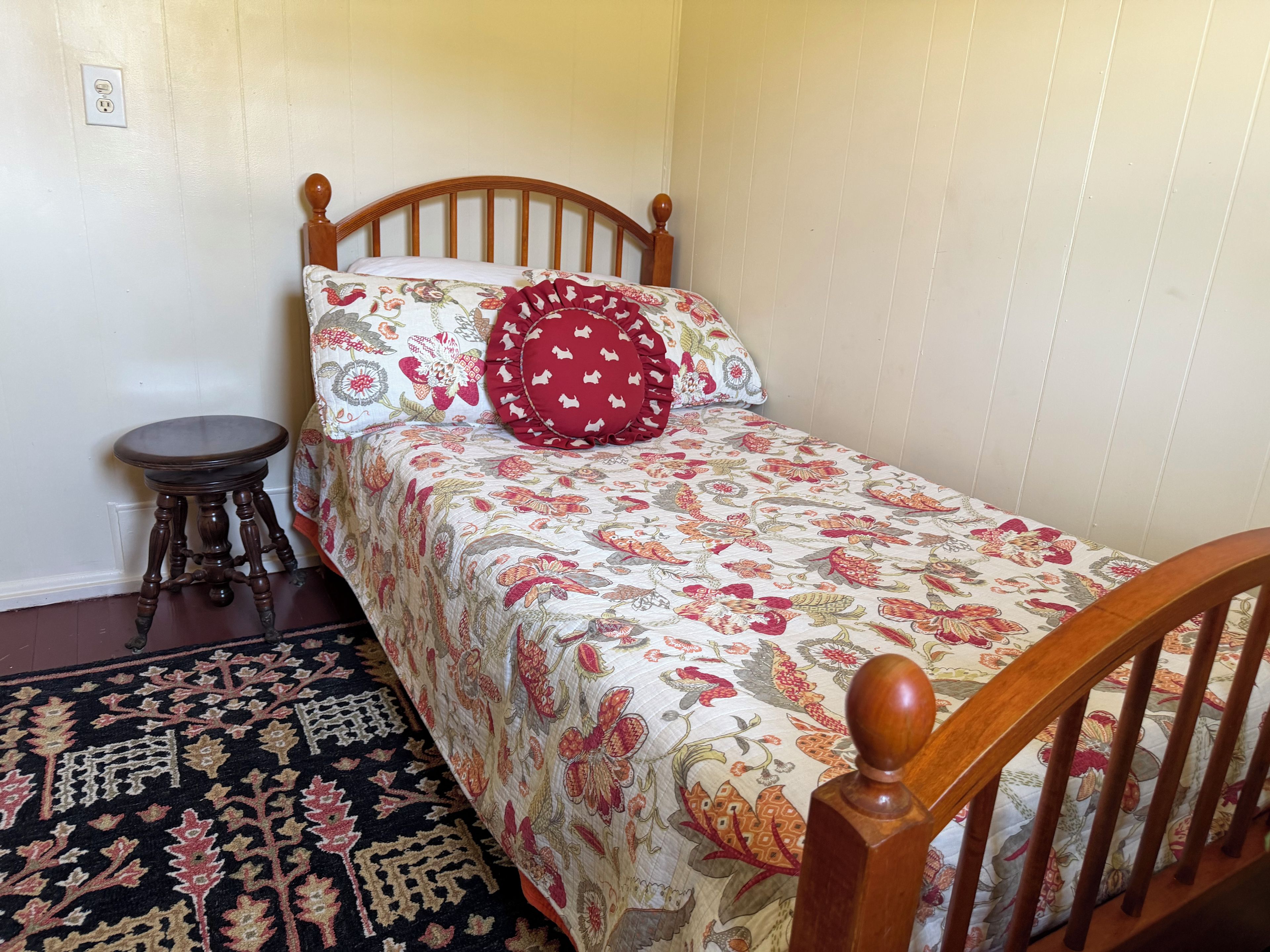 Una habitación pequeña con una cama individual, cabecera de madera, colcha estampada con motivos florales, almohada roja, una mesita redonda y alfombra con diseño.