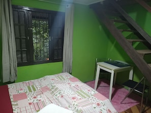 A room with a green wall, a window with a view, a bed with a patterned bedspread, a small white table, and a wooden staircase.