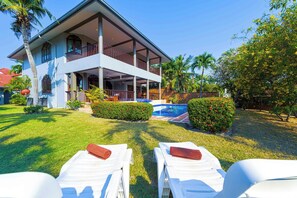 Una casa de dos pisos con balcón, piscina y dos sillones de descanso con toallas.