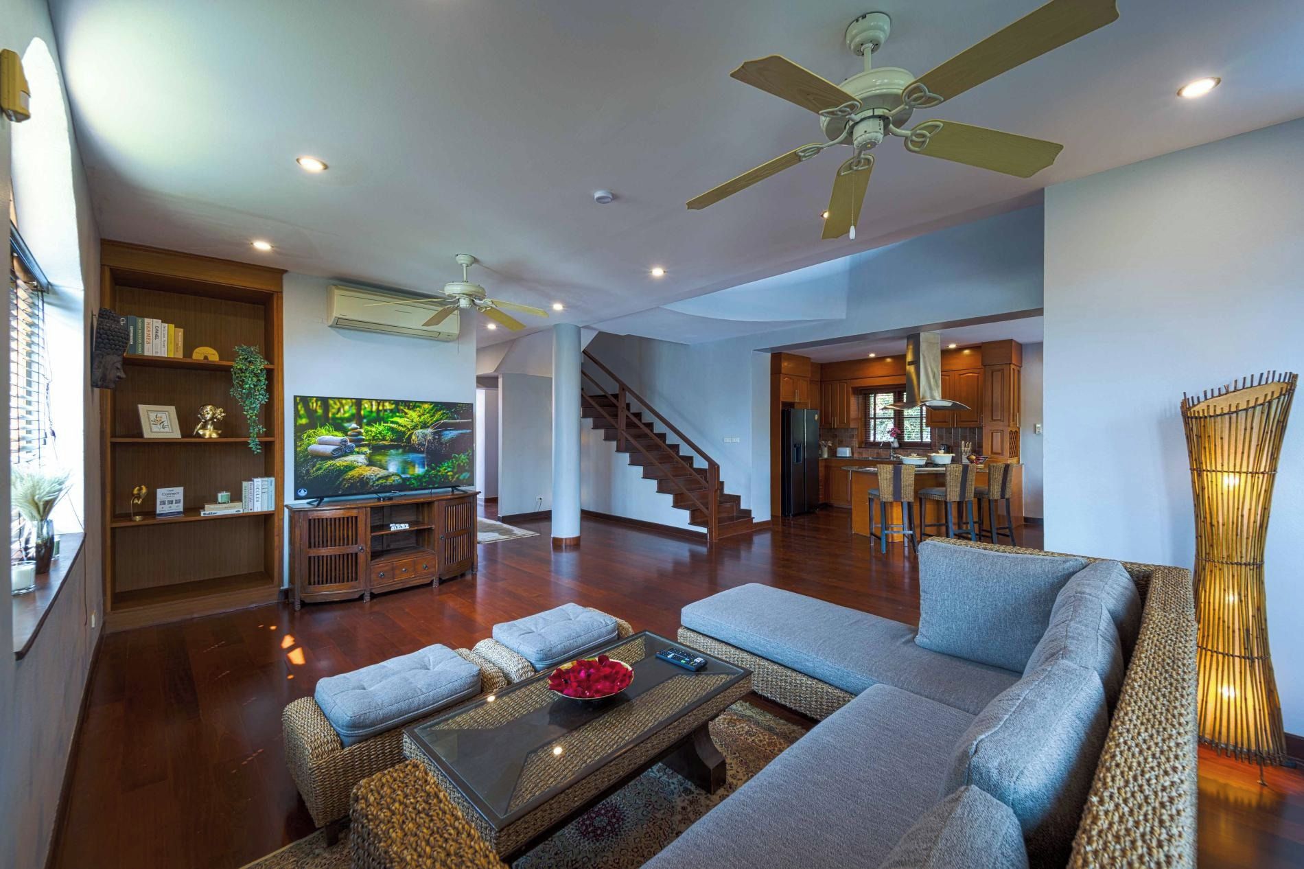 A spacious living area with a sofa, coffee table, and a television on a wooden stand. There is a staircase leading to another room, and a kitchen area is visible in the background.