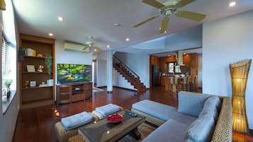 A spacious living area with a sofa, coffee table, and a television on a wooden stand. There is a staircase leading to another room, and a kitchen area is visible in the background.