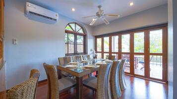 A dining area with a long table set for six, surrounded by wicker chairs, with an air conditioning unit above and a ceiling fan above the table.