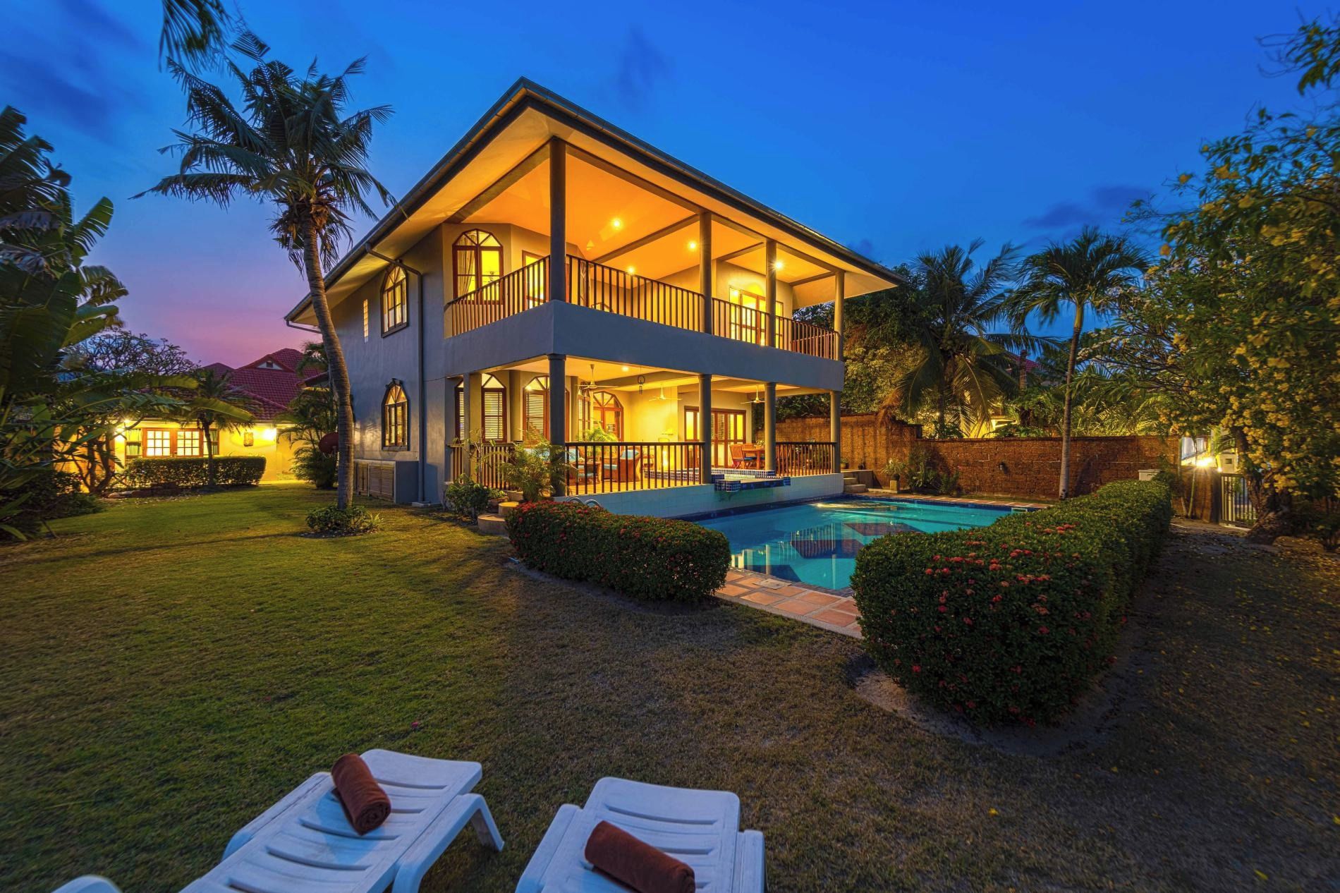Una casa de dos pisos con piscina, rodeada de césped cuidado y plantas tropicales.