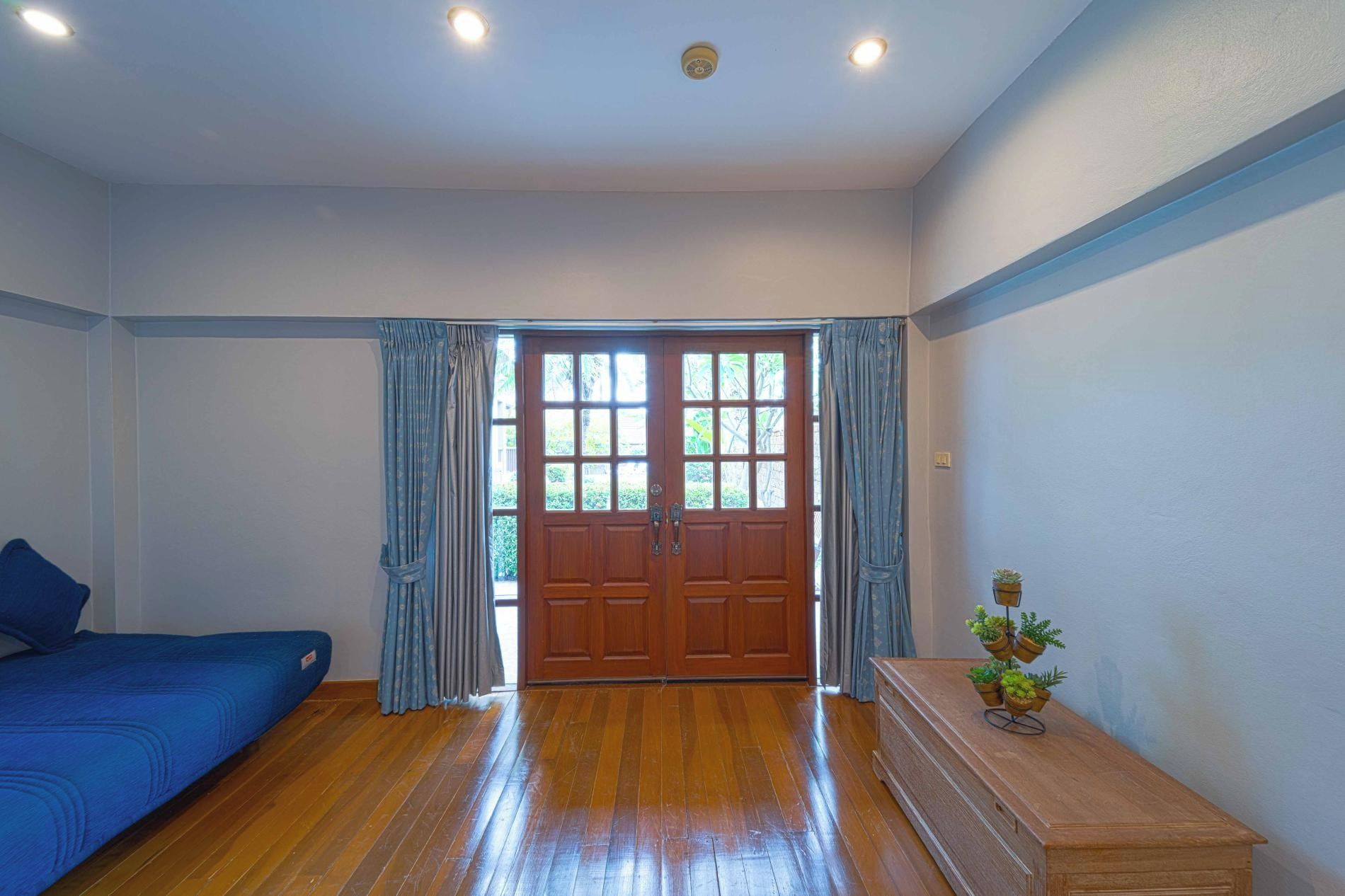 Una habitación con piso de madera, un sofá azul, un armario de madera con plantas y un doble ventanal.