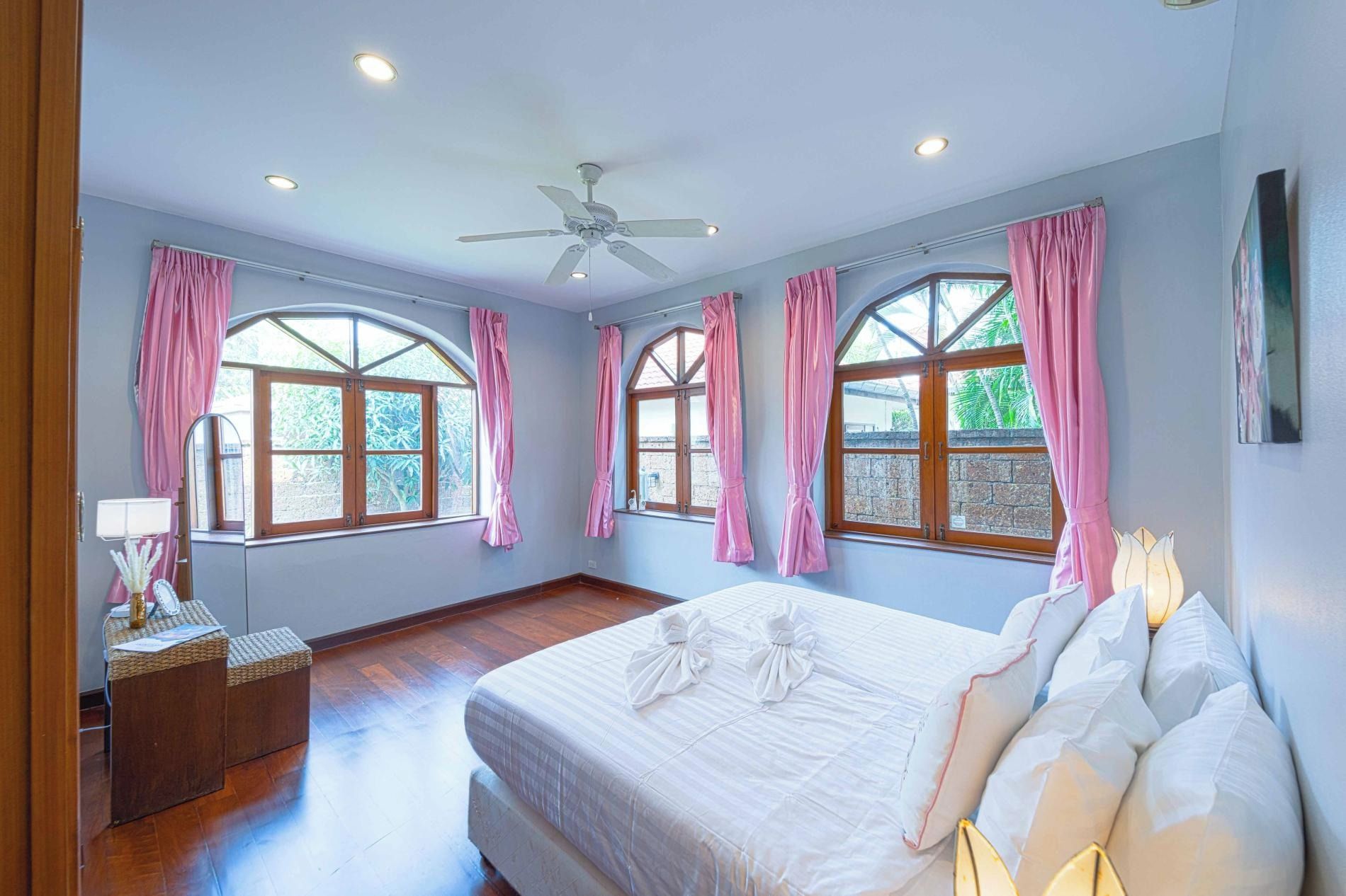 A bedroom with a large bed, two windows with pink curtains, a ceiling fan, and a wooden floor.