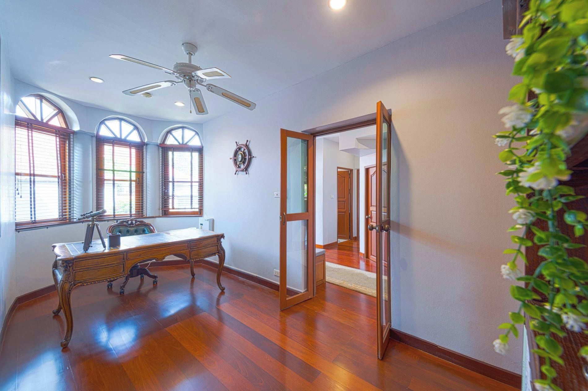 Um quarto espaçoso com uma escrivaninha de madeira, um ventilador de teto e grandes janelas.