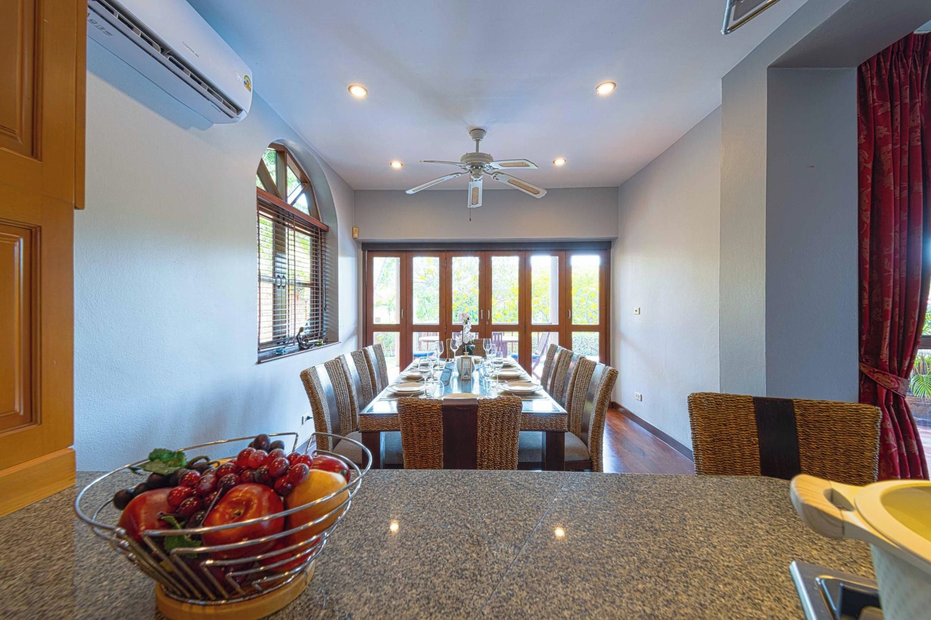 Uma sala de jantar com uma mesa comprida, cadeiras, ventilador de teto e uma janela com persianas.