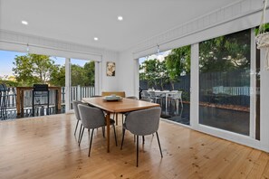 Un vaste espace de salle à manger avec une table en bois et des chaises grises, de grandes portes-fenêtres coulissantes donnant sur un jardin, et une vue sur des arbres et une clôture.