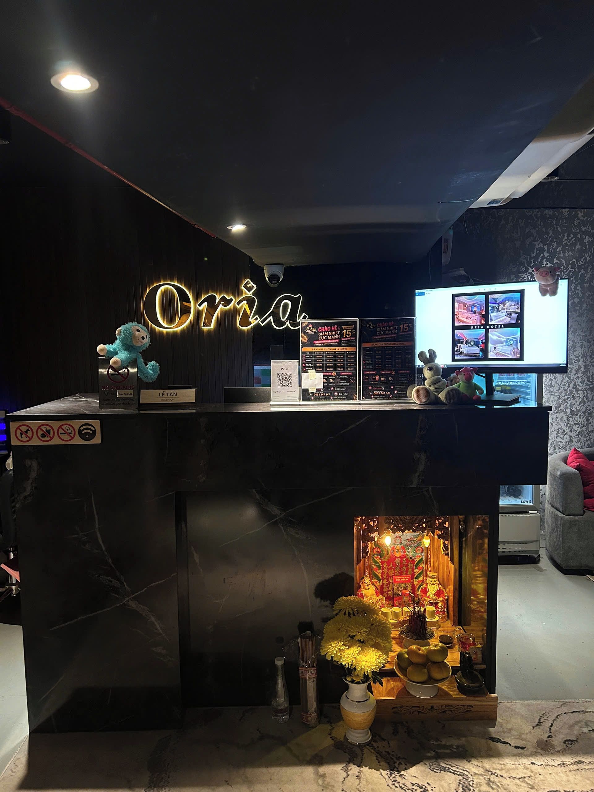 En hotellreception med en neonskylt, en digital skärm som visar hotellinformation och ett litet altare med offergåvor.