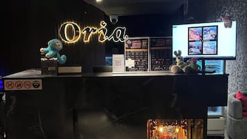 En hotellreception med en neonskylt, en digital skärm som visar hotellinformation och ett litet altare med offergåvor.