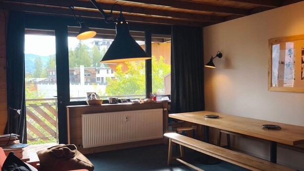 Een knus vertrek met een houten plafond, een bank, een tafel en een raam met uitzicht op gebouwen en bomen.