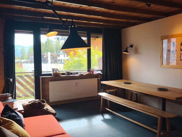 Una habitación acogedora con techo de madera, un banco, una mesa y un ventanal con vistas a edificios y árboles.