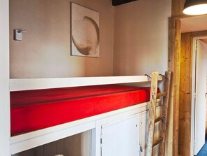 Una cama individual superpuesta con colchón rojo, una escalera de madera y una puerta a la derecha.
