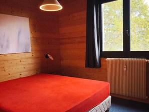 Una habitación con cama, radiador, ventana con cortinas y una lámpara fijada a la pared.