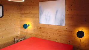Een kamer met houten wanden, een bed met een rode spreidek, een witte plafondlamp en twee wandlampen.