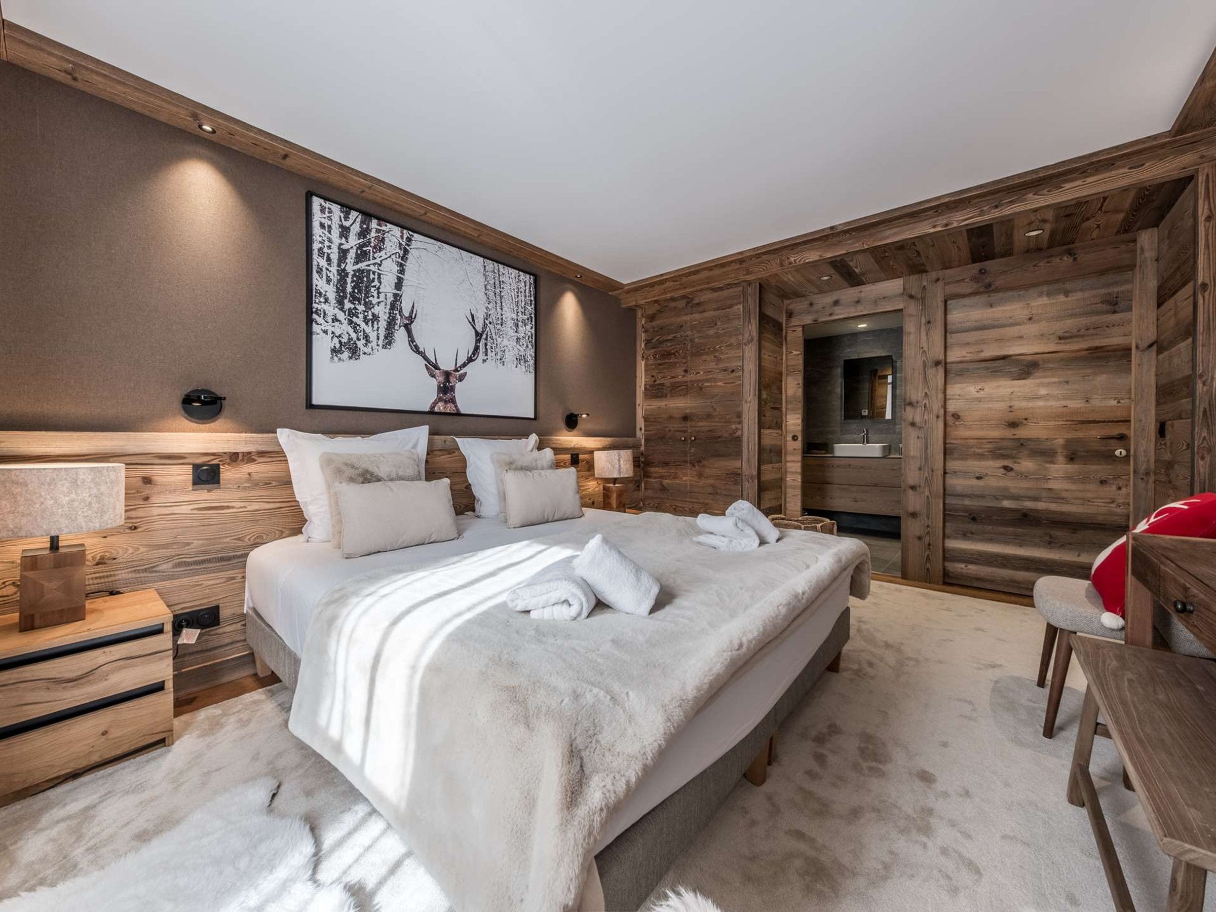A modern bedroom with a large bed, wooden headboard, a wall-mounted artwork, and a visible bathroom door.