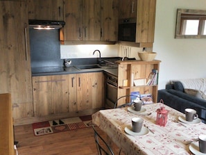 A compact kitchen with wooden cabinets, a microwave, and a sink. There is a small dining area with a table set for two, and a bookshelf with books and a bowl. - Charming apartment in the village - 8 guests (Arêches-Beaufort)