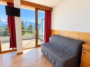 A room with a wooden floor, a grey sofa, a wooden cabinet, a television, and a sliding glass door with red curtains. - Apartment with south-facing terrace - 6 guests (Chamrousse)